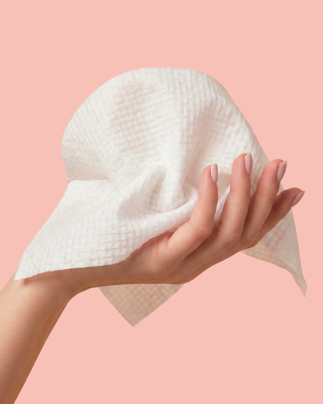 A hand with manicured nails holds a white textured waffle-weave facial cleansing cloth against a solid pastel pink background.
