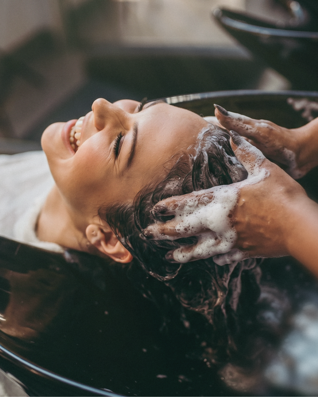 A person lying back in a salon shampoo bowl with dark, soapy hair being washed by another person's hands.
