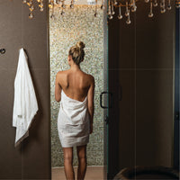 Image of a Scrummi Original Waffle Body Towel in white in use within a spa changing room. The towel is depicted in action, perhaps wrapped around a client and hanging on a hook, showcasing its soft, waffle-textured material. The disposable and biodegradab