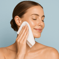 Smiling woman gently cleansing her face with a soft white facial towel against a light blue background.