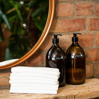 Two brown bottles with black pumps on a wooden shelf with white waffle scrummi towels against a brick wall.